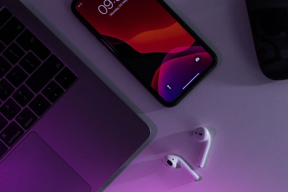 A phone and wireless earbuds on a desk beside a laptop