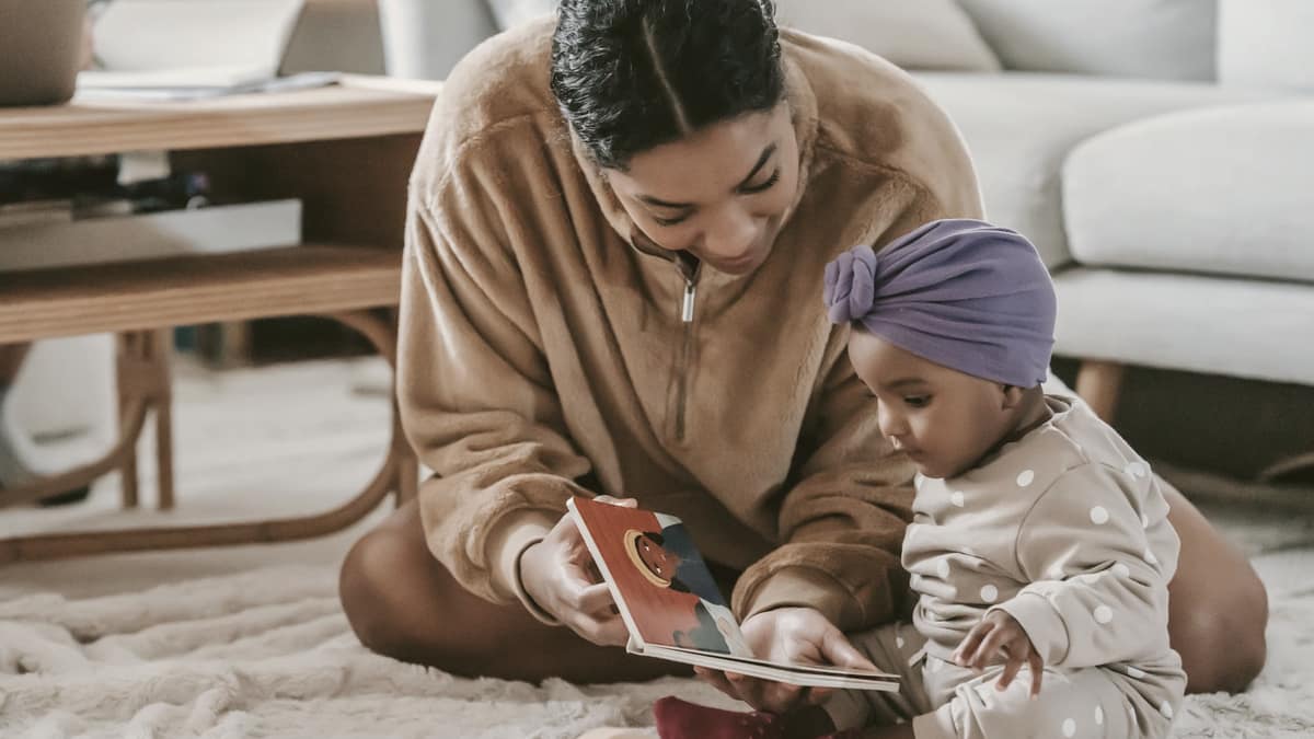A parent sitting on the floor showing a picture book to a baby