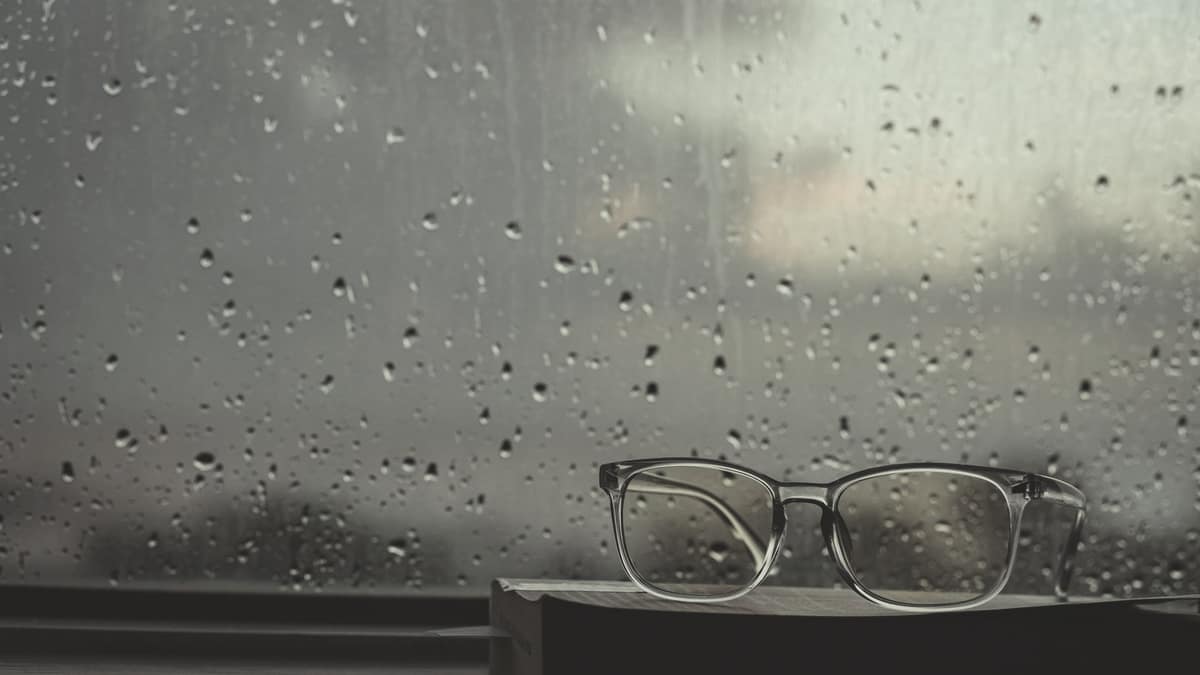 Reading glasses resting on a book beside a rain-streaked window
