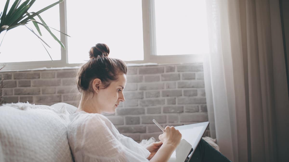 A woman reading on a tablet on a sofa beside a window