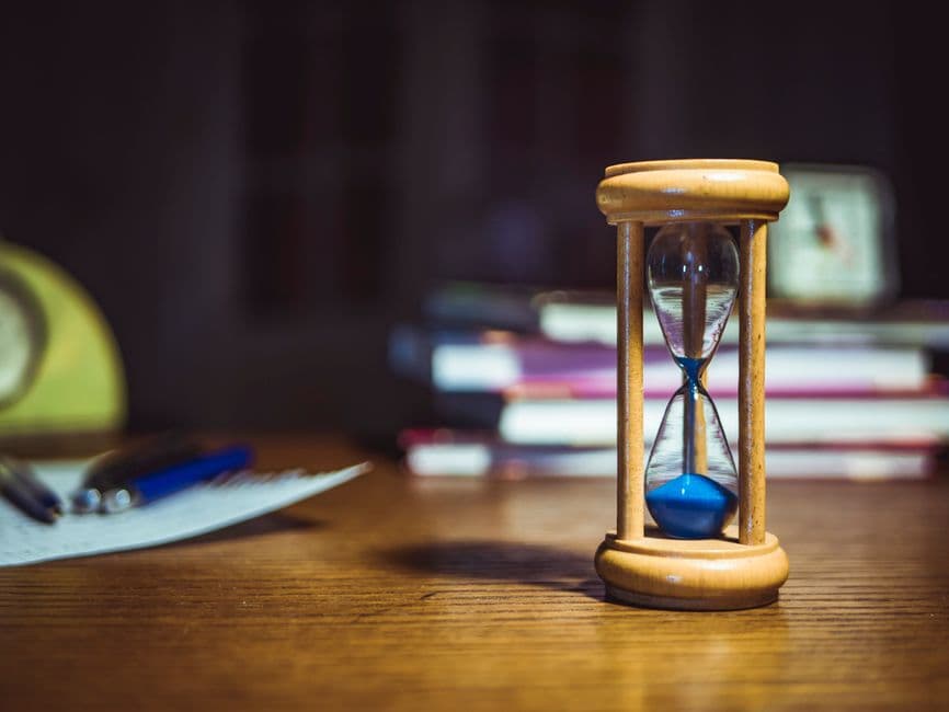 A wooden hourglass on a desk beside books and pens