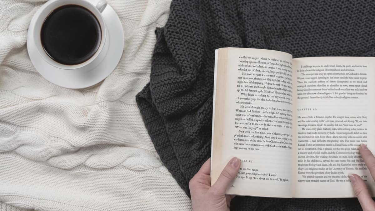 Hands holding an open book beside a cup of coffee on a blanket