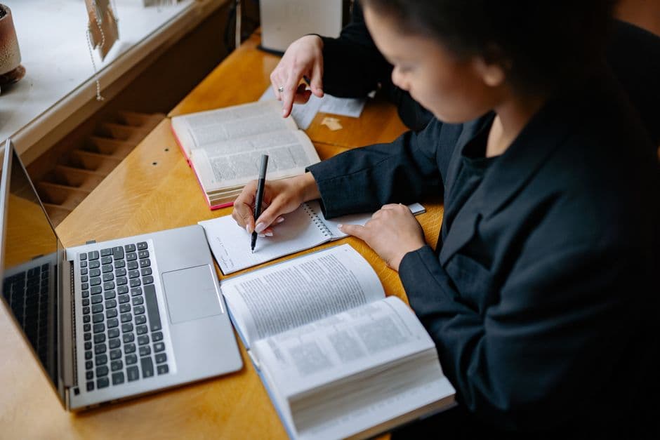 Two students taking notes beside books and a laptop