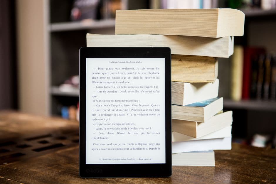 An e-reader beside a stack of physical books on a wooden table